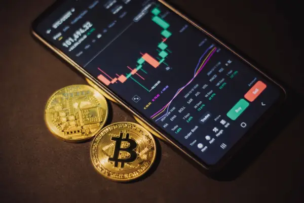 Smartphone showing a crypto trading app next to two bitcoin coins